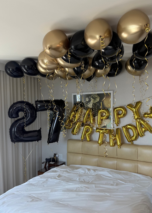 A hotel room bedroom decorated for a 27th birthday celebration with a balloon garland in black and metallic gold, a large black number 27 balloon, and gold foil 'HAPPY BIRTHDAY' balloons above a bed with white bedding.
