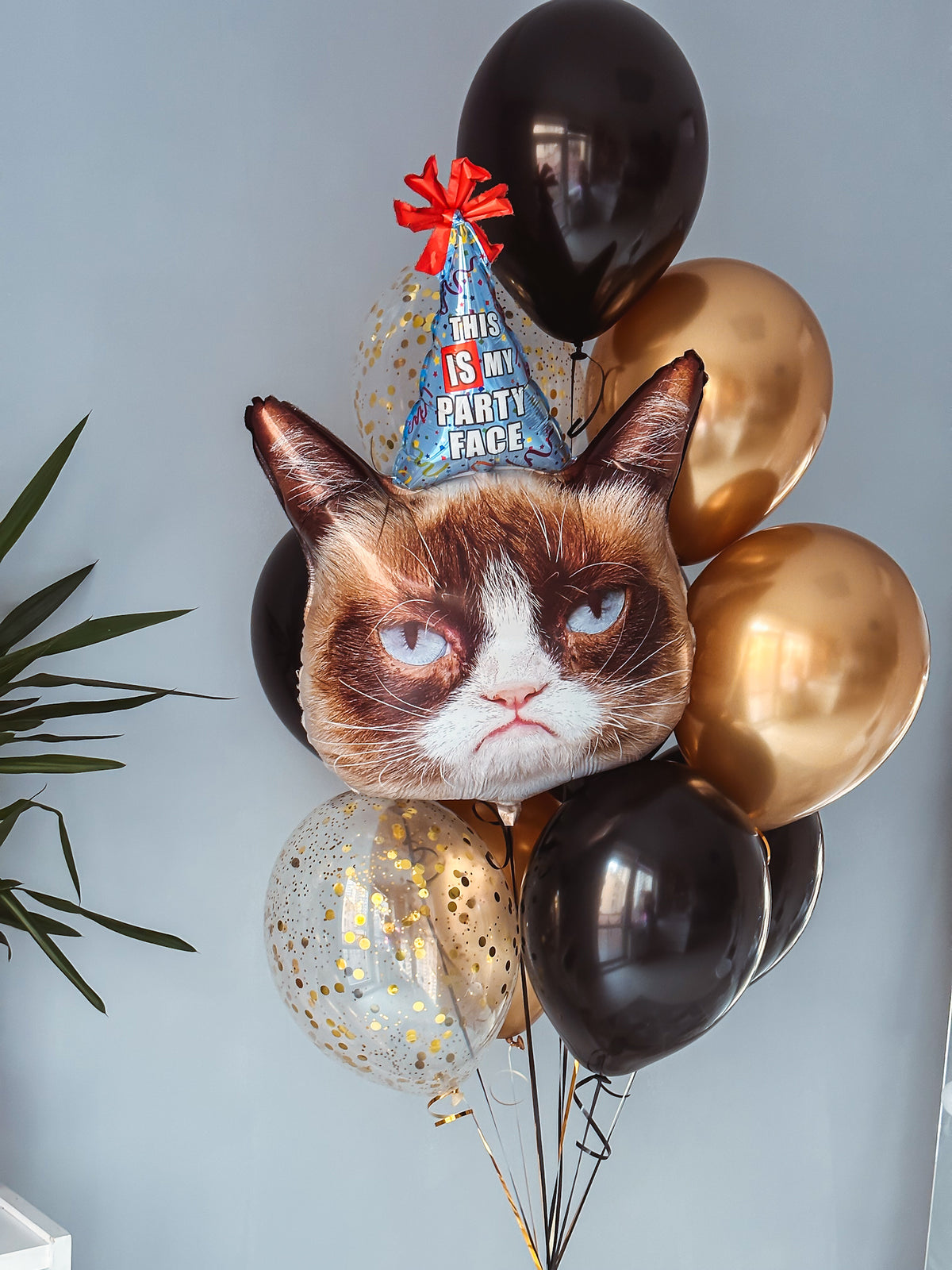 Funny cat-shaped birthday balloon with a party hat, surrounded by black, metallic gold, and gold confetti helium balloons.