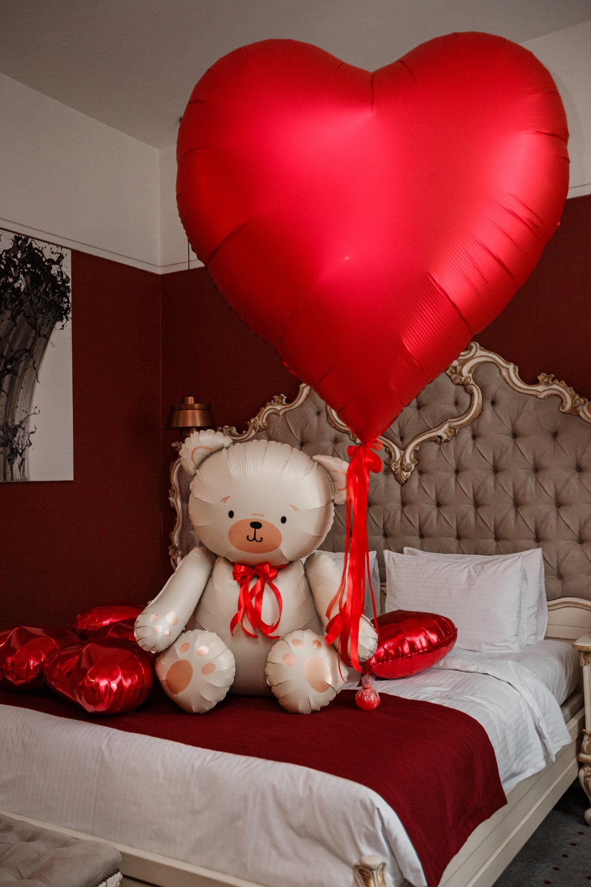 Romantic bedroom balloon decoration featuring an oversized satin-finish red heart helium balloon with flowing red ribbons, a large cream teddy bear foil balloon with a red bow, and coordinating metallic red foil heart balloons. Valentine's Day and anniversary surprise styling by Best Balloons LA.