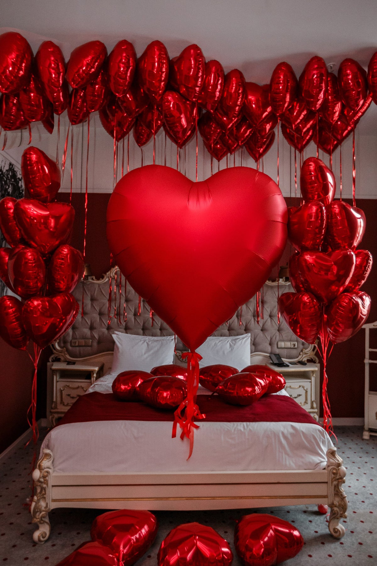 Romantic bedroom decoration featuring a giant satin red heart helium balloon, metallic red foil heart balloons floating across the ceiling with long red ribbons, coordinating red heart balloon bouquets on both sides of the bed, and scattered red foil hearts. Luxury Valentine's Day, anniversary, and proposal balloon decoration by Best Balloons LA.