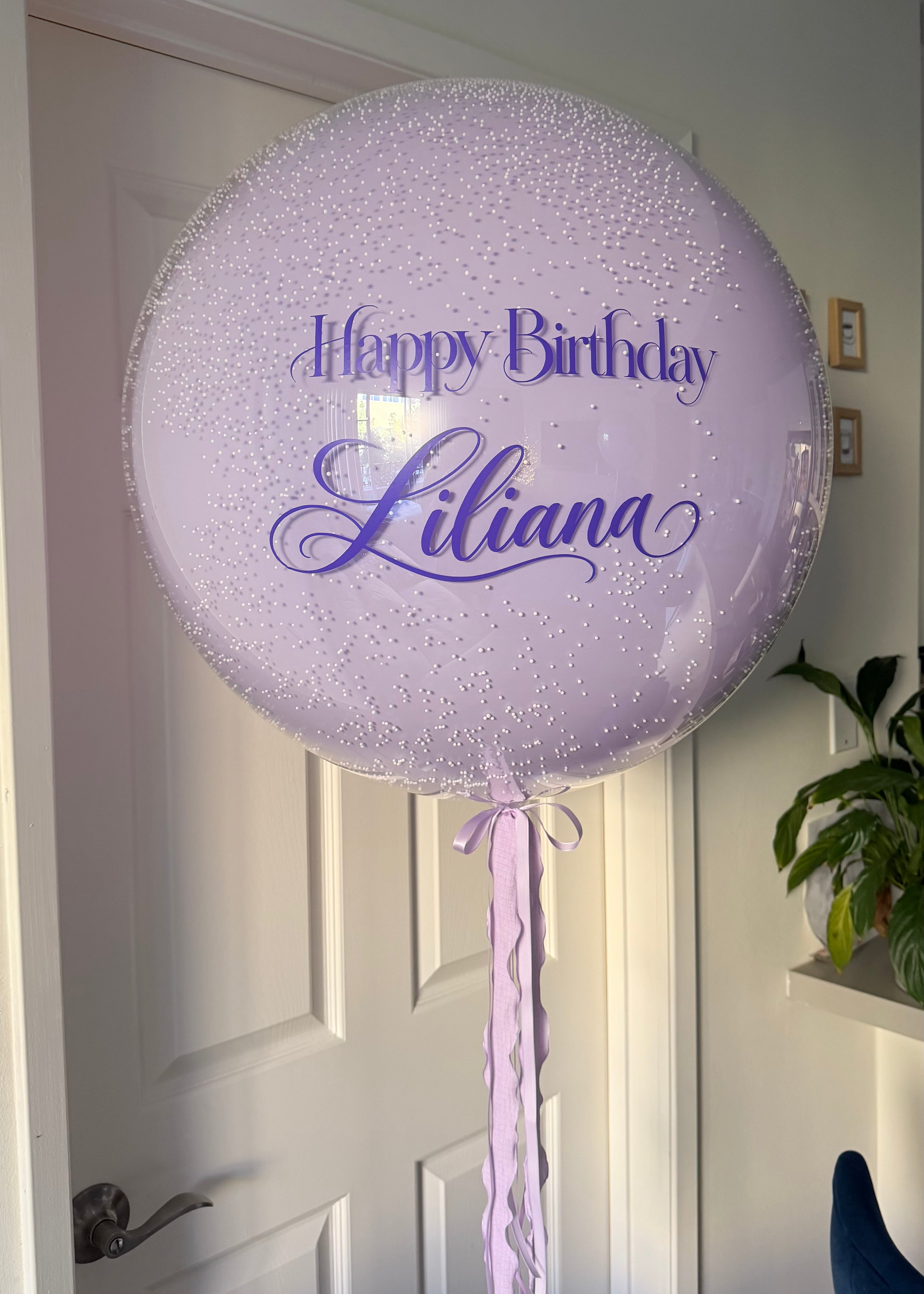 Elegant transparent bubble balloon featuring a soft lavender inner balloon, delicate white confetti-like accents, purple Happy Birthday Liliana lettering, and long flowing lavender ribbons. Personalized helium birthday balloon by Best Balloons LA.