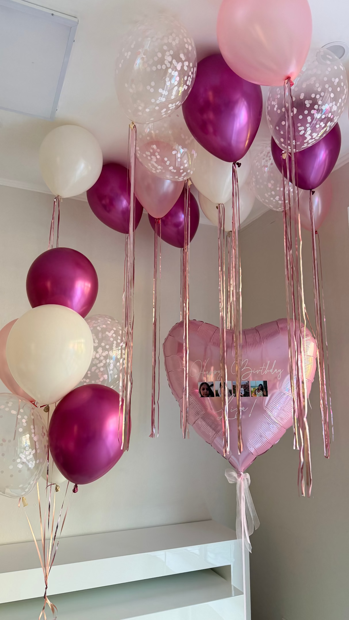Pink, fuchsia, white, and confetti helium balloons with long metallic ribbons and a large personalized pink heart balloon for a birthday room setup.