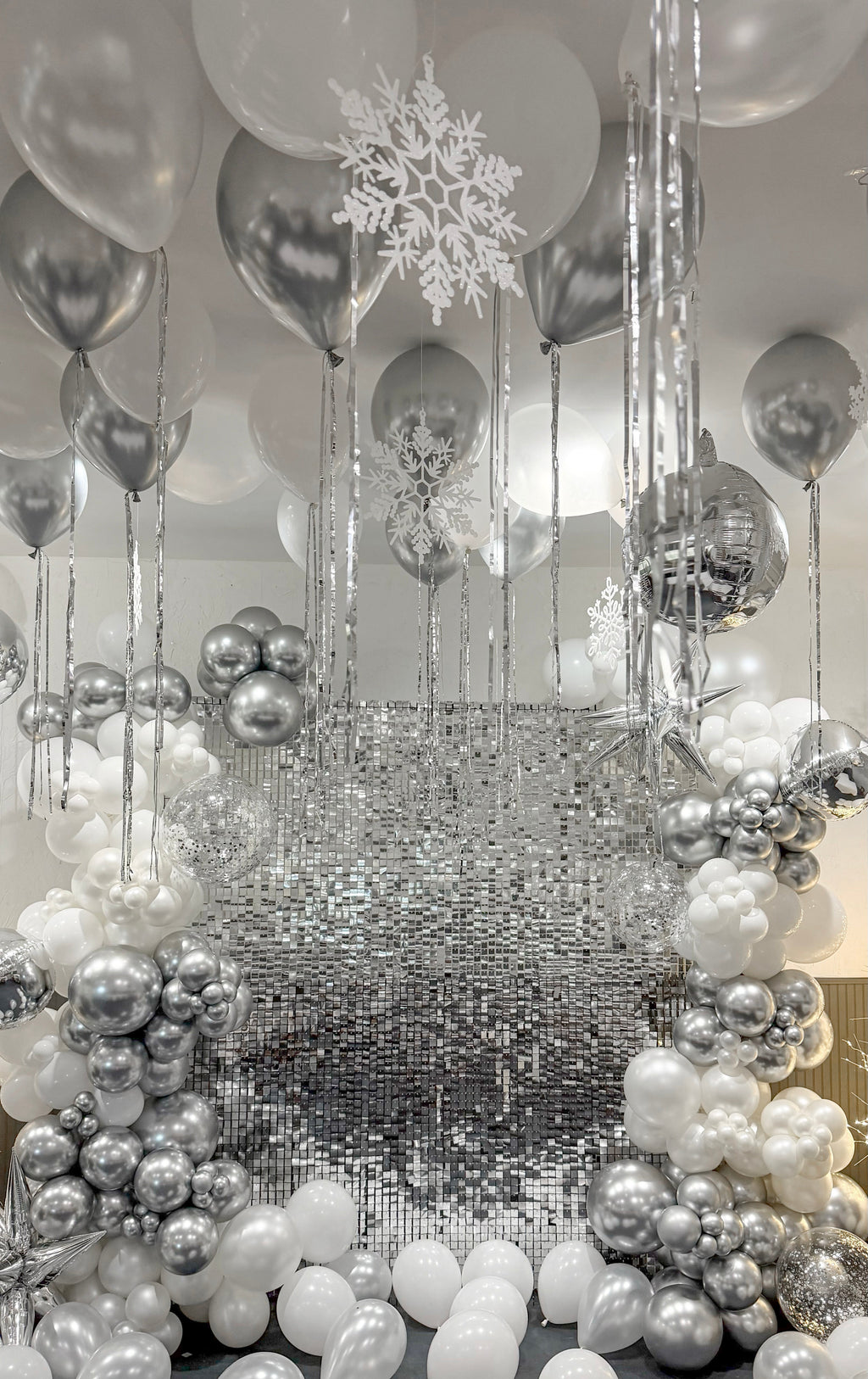 Create a spectacular Winter Wonderland Balloon Backdrop with a glamorous mix of silver, white, chrome, and clear balloons surrounding a shimmering sequin wall.