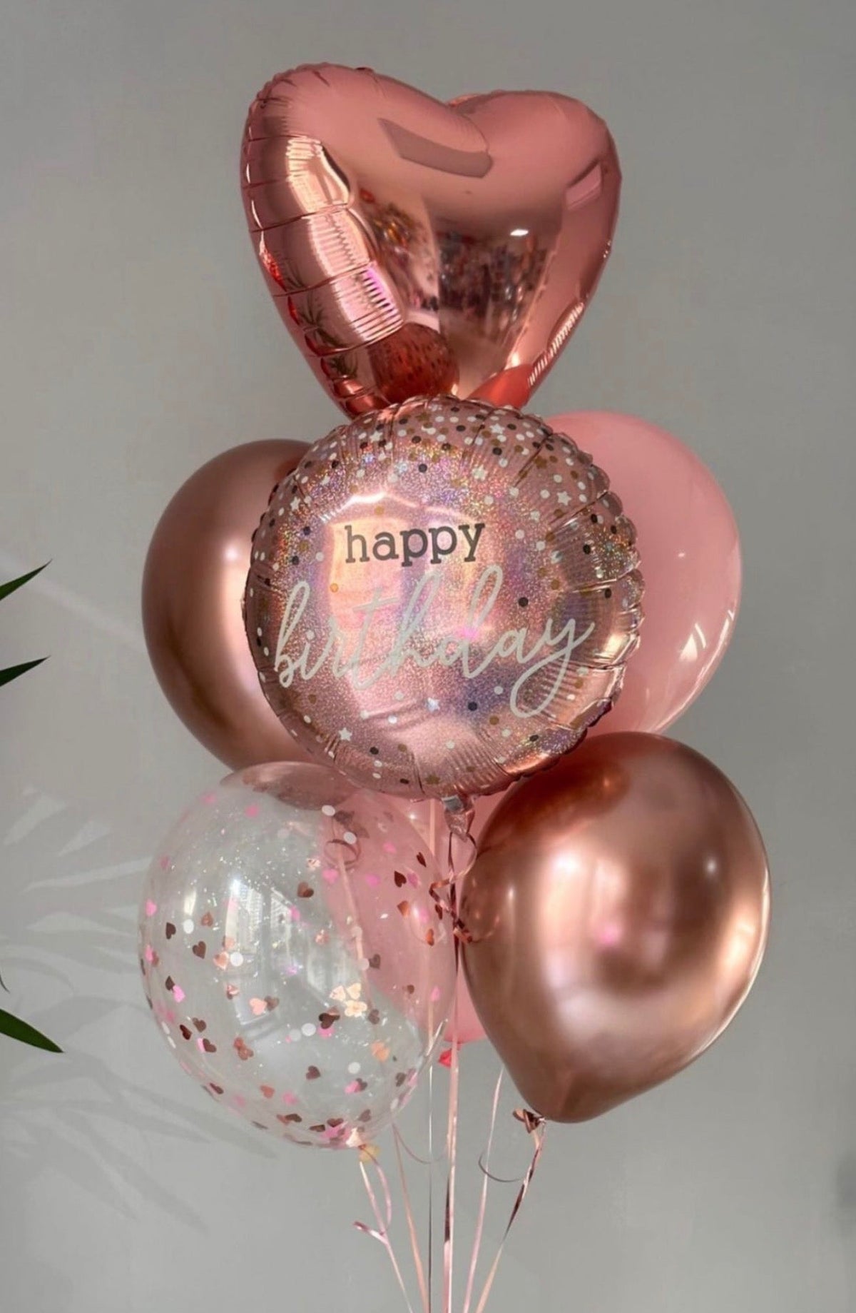A cluster of metallic and clear birthday balloons in rose gold, pink, and clear with confetti, featuring a heart-shaped balloon and a round clear balloon printed with 'happy birthday'.