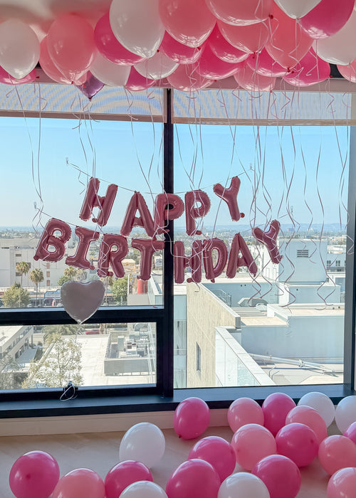 Pink and white balloon decorations arranged around a large window, with pink foil letter balloons spelling 'HAPPY BIRTHDAY' and a small heart balloon hanging near the glass.