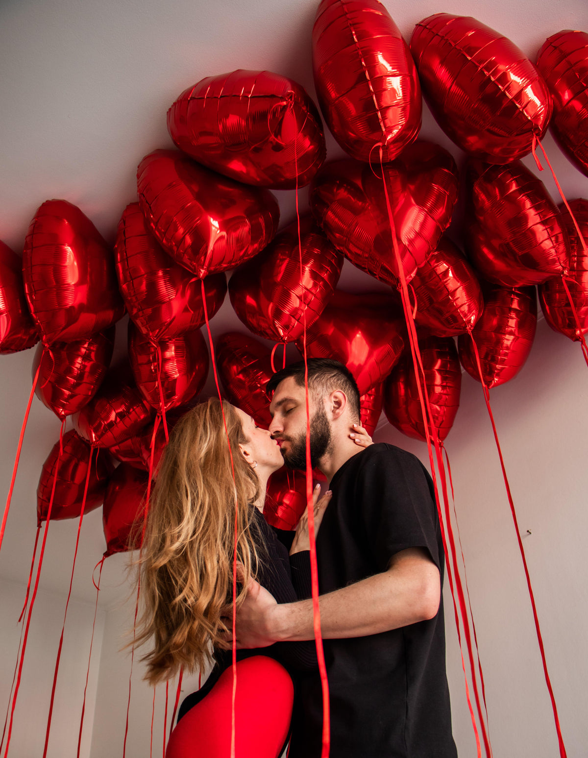 Romantic indoor decoration with floating metallic red heart-shaped helium balloons, long red ribbons, and a couple sharing a kiss. Luxury Valentine's Day room decoration, anniversary surprise, and marriage proposal balloon styling by Best Balloons LA in Beverly Hills, Bel Air, Malibu, and Los Angeles.